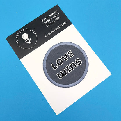 An embroidered felt patch with the text "LOVE WINS" on a display card. The patch is ready for sewing or ironing onto fabric, with the brand "The Unruly Stitch" displayed on the card.