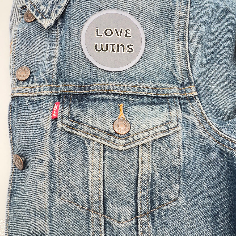 A denim jacket with a "LOVE WINS" embroidered patch sewn on the upper pocket area. The patch features white text on a black background with a gray border.