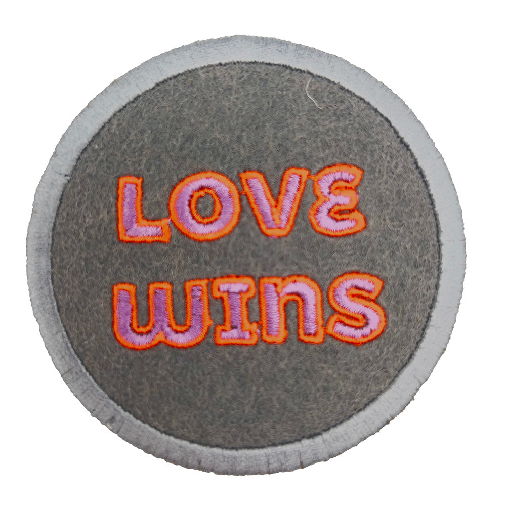 A close-up image of a round embroidered felt patch with the text "LOVE WINS" in pink letters outlined in orange, set against a gray background with a light gray border.