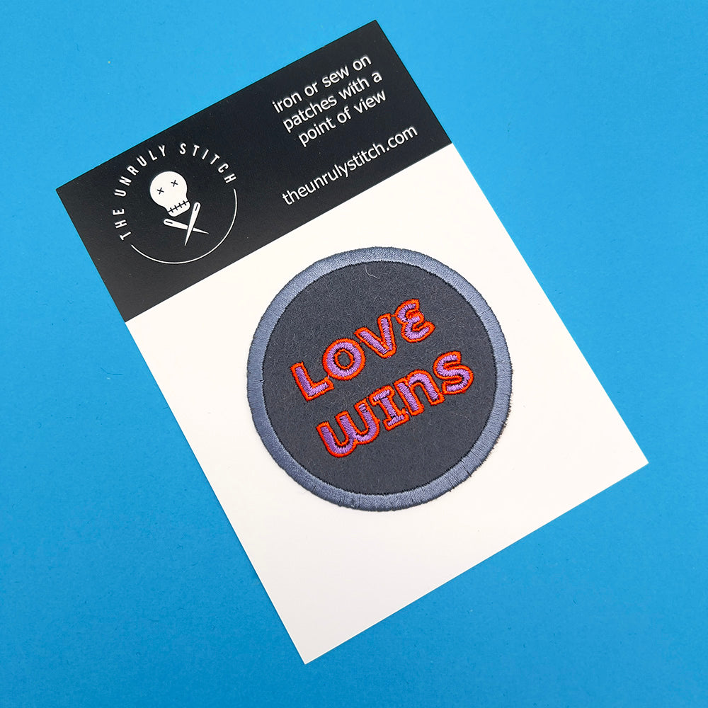 An embroidered felt patch with the text "LOVE WINS" in pink and orange letters on a display card. The patch is ready for sewing or ironing onto fabric, with the brand "The Unruly Stitch" displayed on the card.