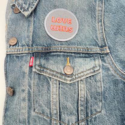 A denim jacket with a "LOVE WINS" embroidered patch sewn on the upper pocket area. The patch features pink text outlined in orange on a gray background with a light gray border.