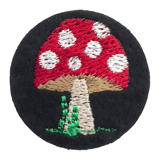 Close-up of a felt badge with an embroidered red and white spotted mushroom on a black background, designed by The Unruly Stitch.