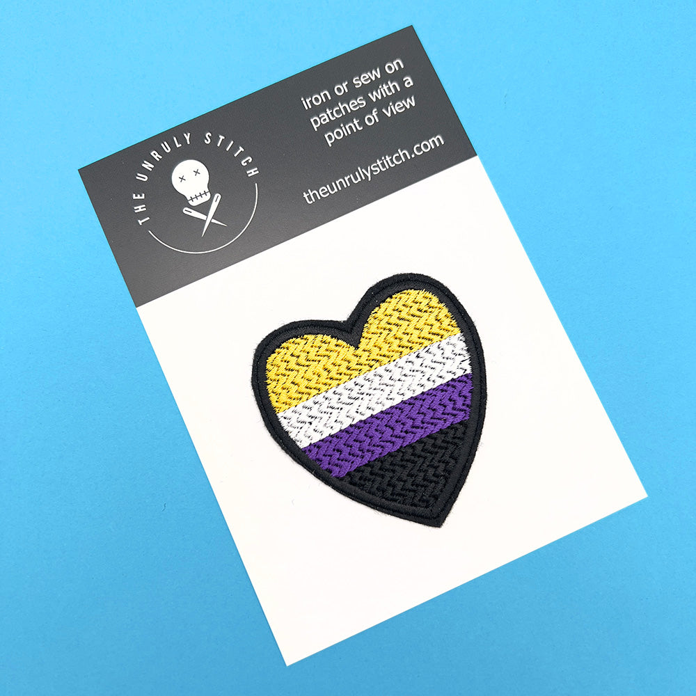 A heart-shaped embroidered felt patch with the non-binary pride flag colors attached to a card. The card has a black header with the brand's logo, "The Unruly Stitch," and the website address.