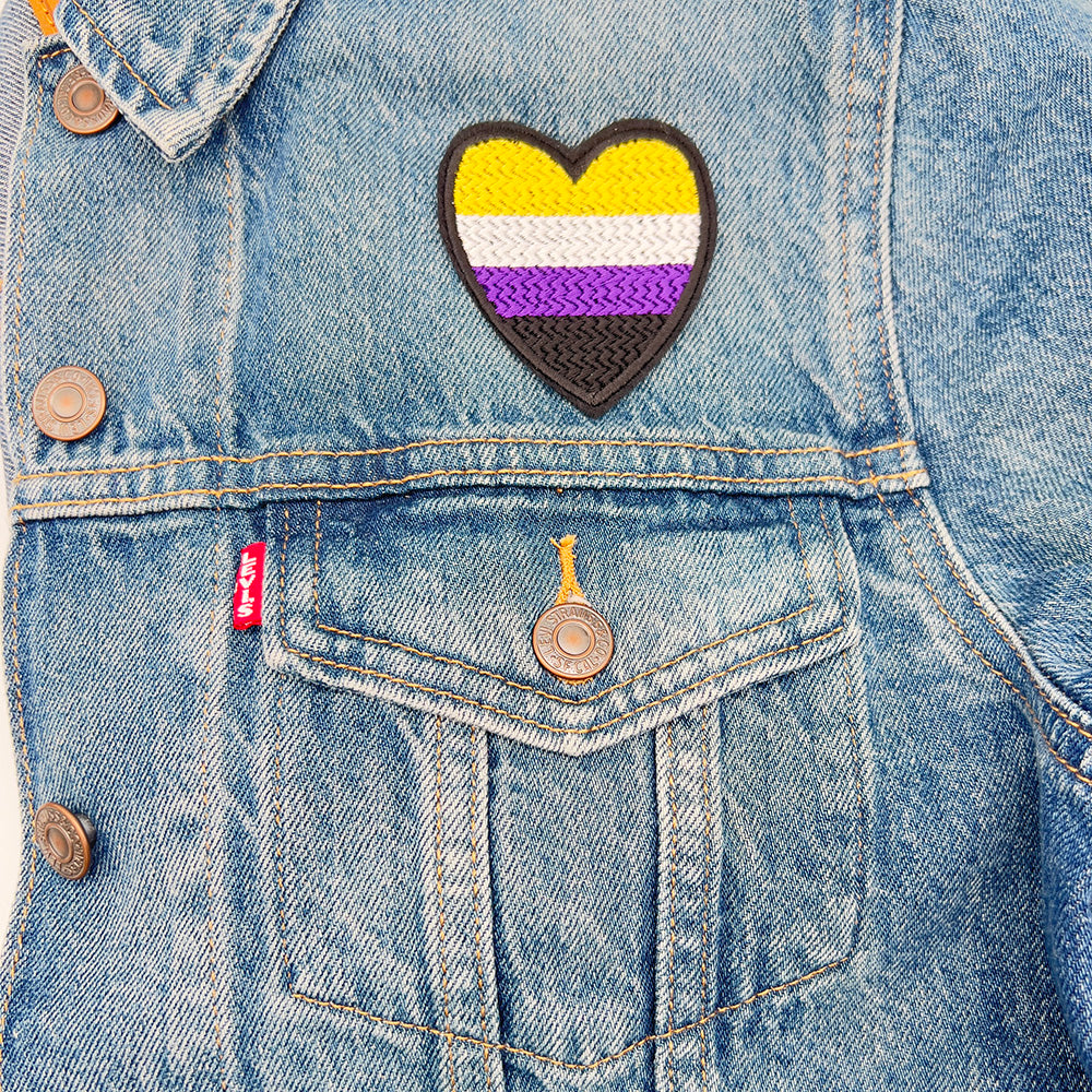 A heart-shaped embroidered felt patch with the non-binary pride flag colors affixed to the upper left part of a denim jacket.