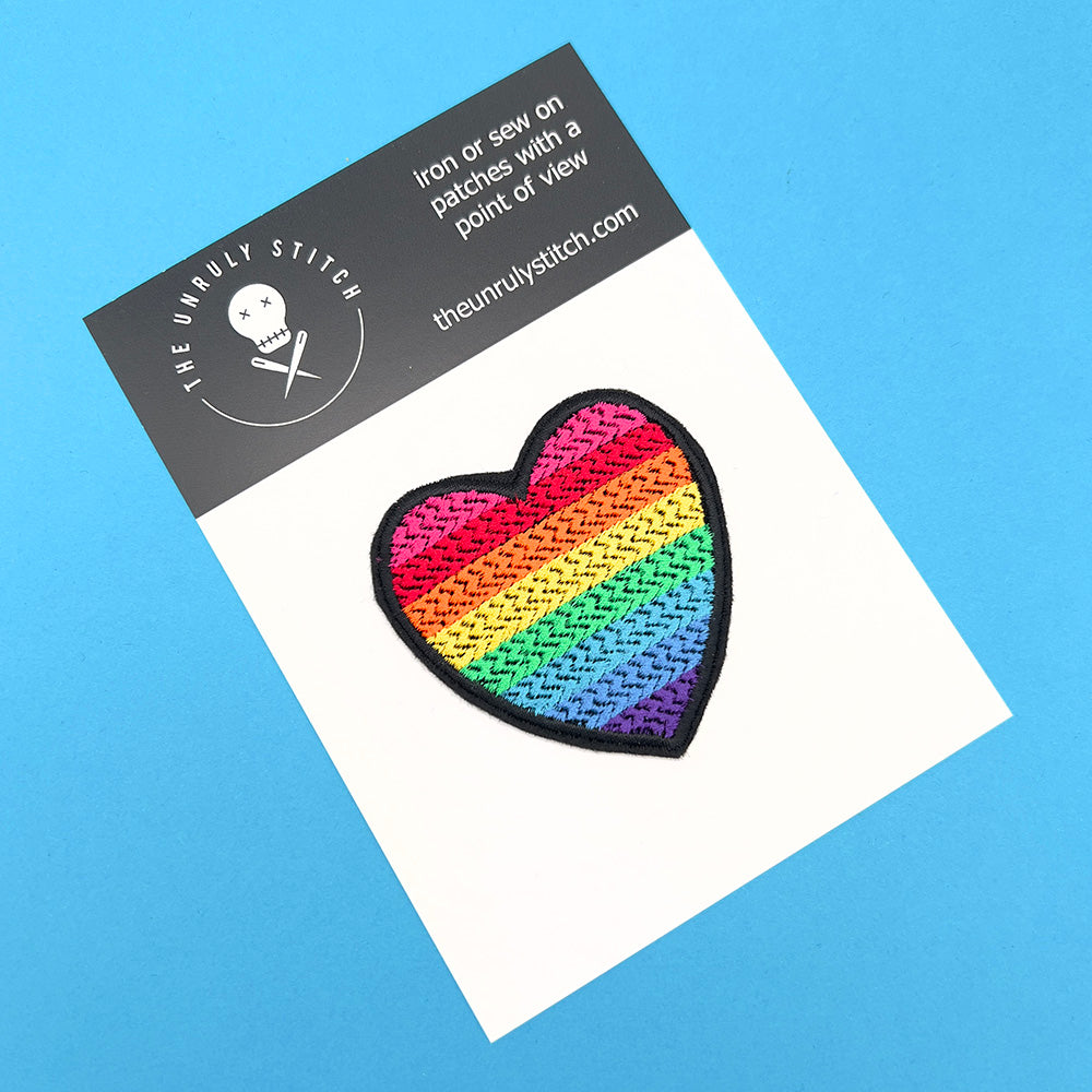 A heart-shaped embroidered patch with the rainbow colors of the original gay pride flag, mounted on a branded card from The Unruly Stitch.