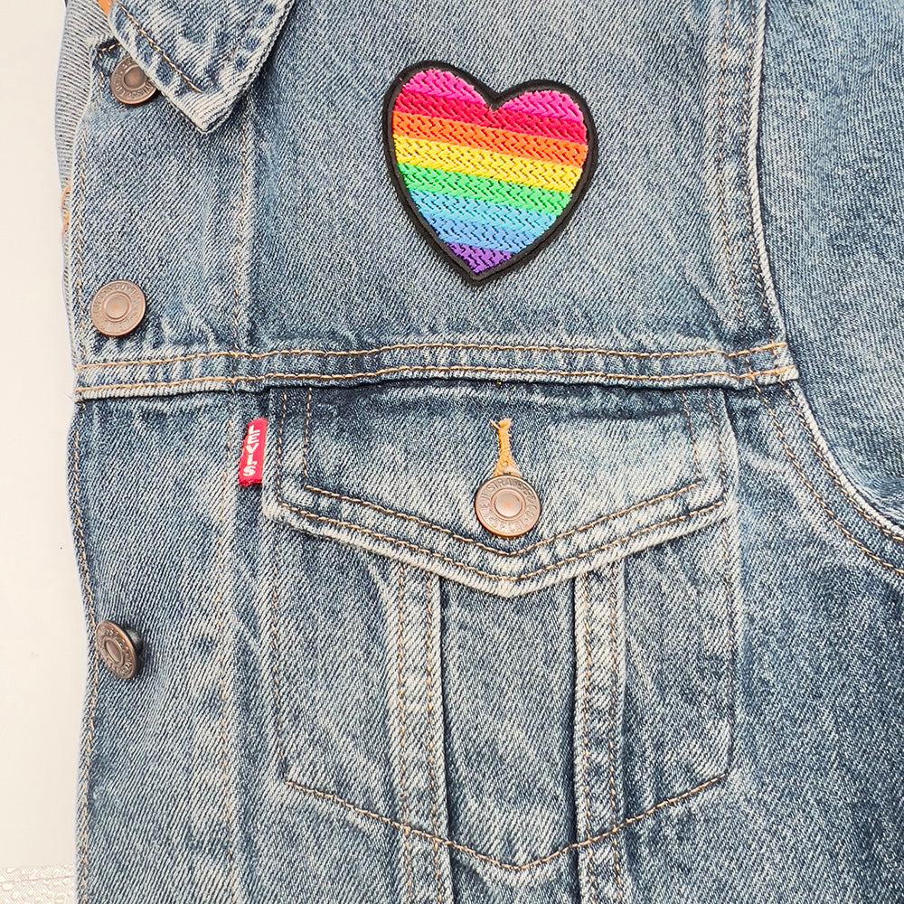 A heart-shaped embroidered patch with the rainbow colors of the original gay pride flag, ironed above the upper left pocket of a blue denim jacket.