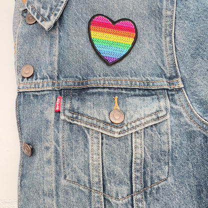 A heart-shaped embroidered patch with the rainbow colors of the original gay pride flag, ironed above the upper left pocket of a blue denim jacket.