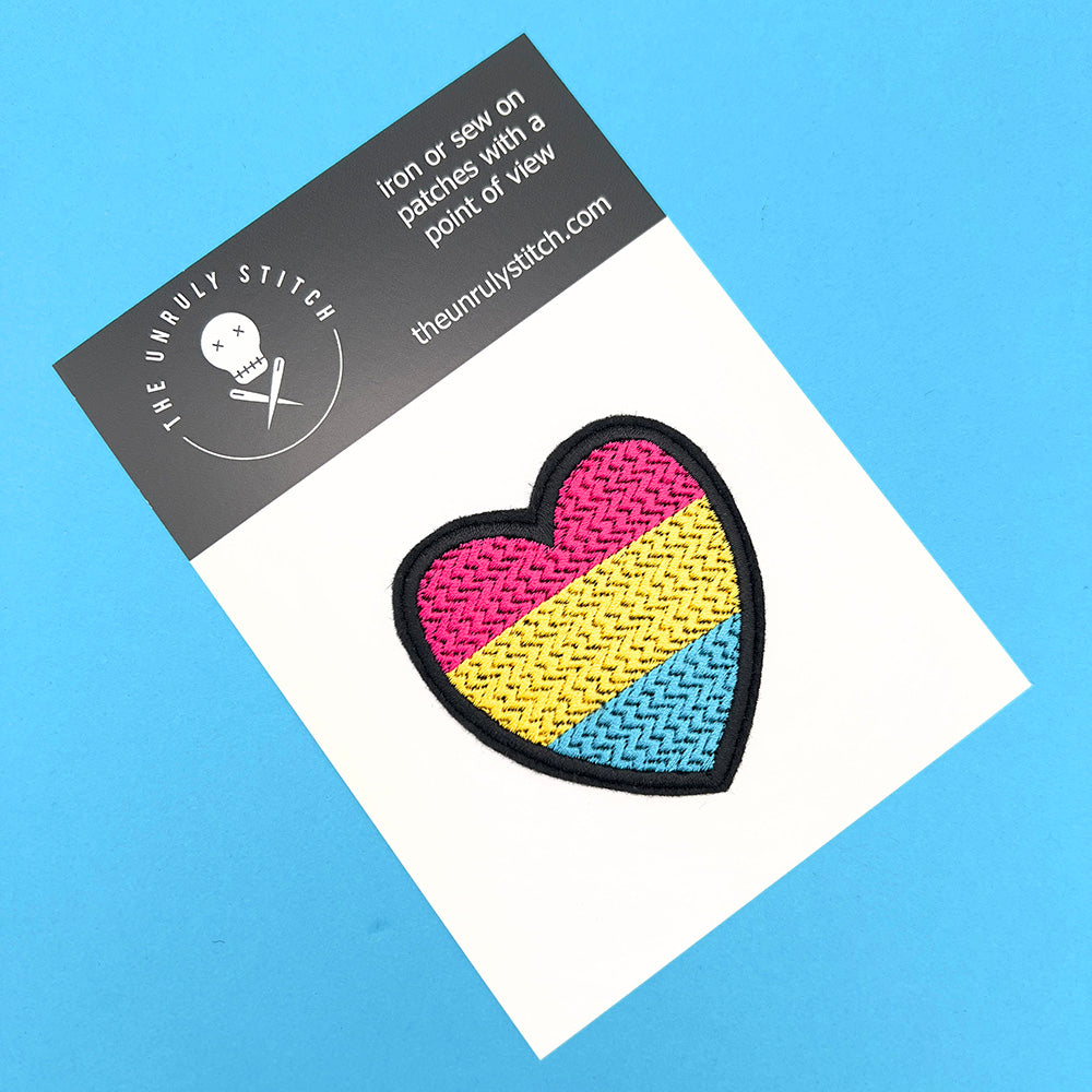 A heart-shaped embroidered patch with the colors of the pansexual pride flag: pink, yellow, and blue, mounted on a branded card from The Unruly Stitch. The card has a white background and sits on a blue surface.