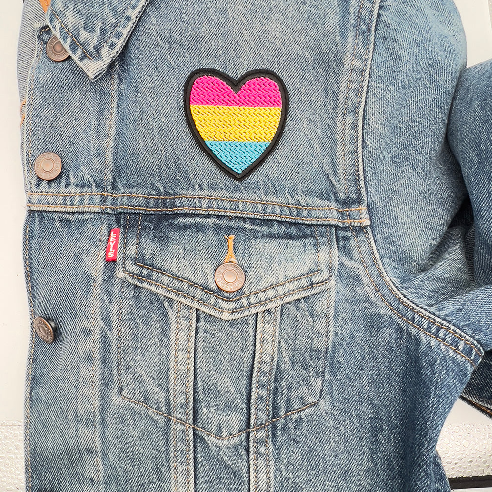 A heart-shaped embroidered patch with the colors of the pansexual pride flag: pink, yellow, and blue, ironed above the upper left pocket of a blue denim jacket.