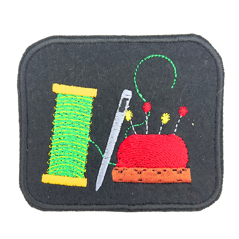 Close-up view of an embroidered felt patch depicting a spool of green thread, a silver needle, and a red pincushion with multicolored pins. The patch is rectangular with a black background and detailed stitching.