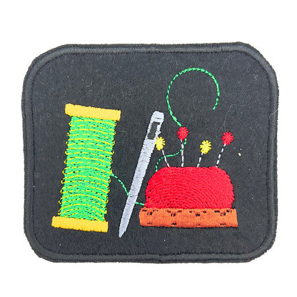 Close-up view of an embroidered felt patch depicting a spool of green thread, a silver needle, and a red pincushion with multicolored pins. The patch is rectangular with a black background and detailed stitching.