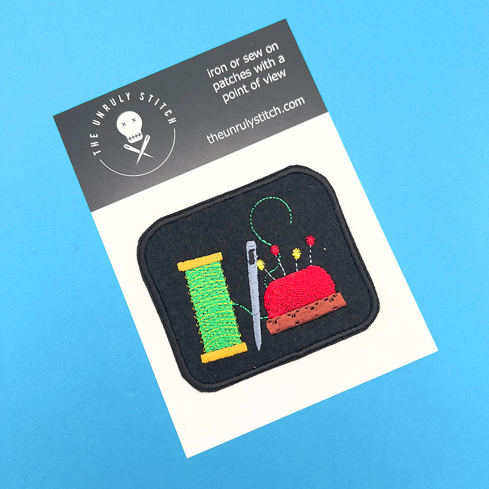 Image of an embroidered felt patch depicting a spool of green thread, a silver needle, and a red pincushion with multicolored pins, displayed on a card with branding. The patch is rectangular with a black background and detailed stitching.