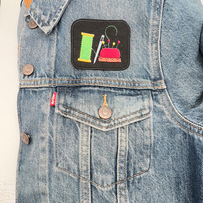 Embroidered felt patch depicting a spool of green thread, a silver needle, and a red pincushion with multicolored pins, sewn onto the pocket area of a blue denim jacket. The patch is rectangular with a black background and detailed stitching.