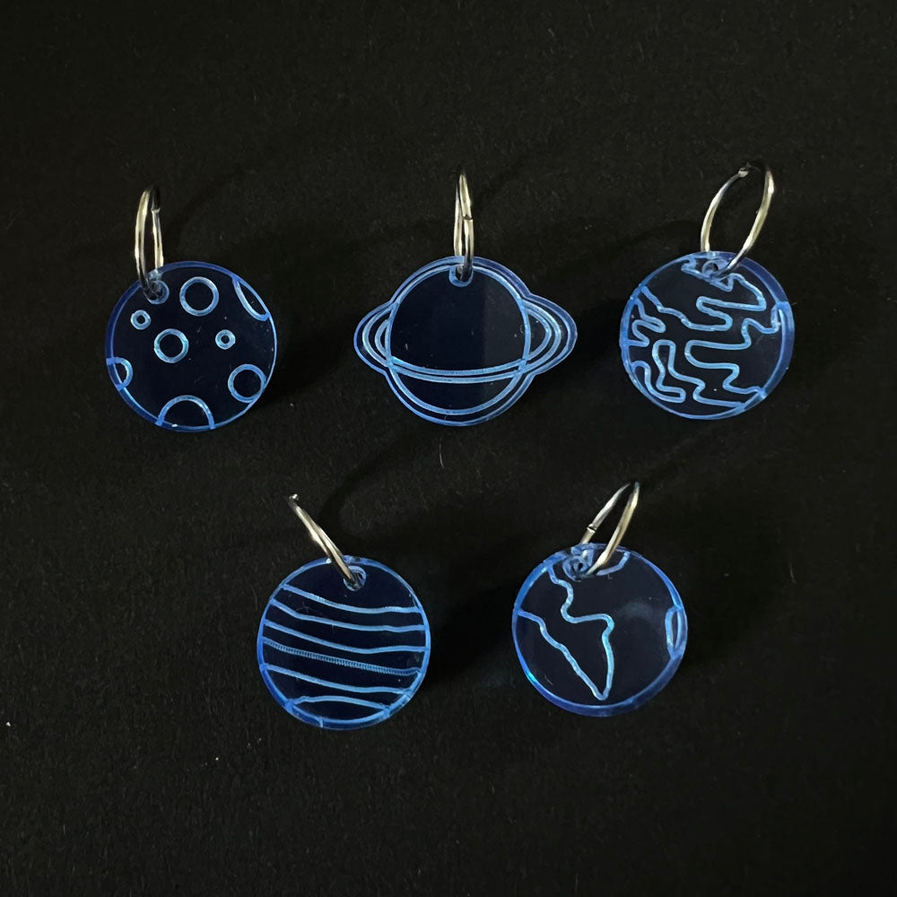 Five blue perspex stitch markers with designs representing different planets and celestial bodies. Each marker is attached to a metal jump ring and arranged on a black background.