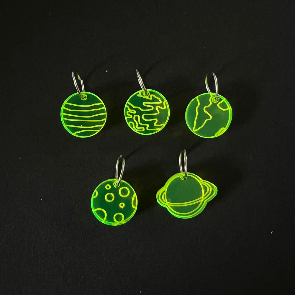 Five green perspex stitch markers featuring various planet designs. Each marker is attached to a metal jump ring and displayed on a black background.