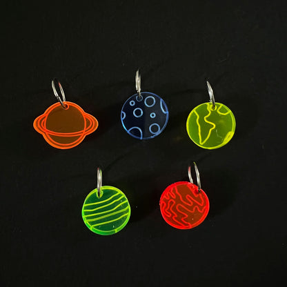 Five perspex stitch markers in various colors (green, blue, red, and orange) depicting different planets. Each marker is attached to a metal jump ring and arranged on a black background.