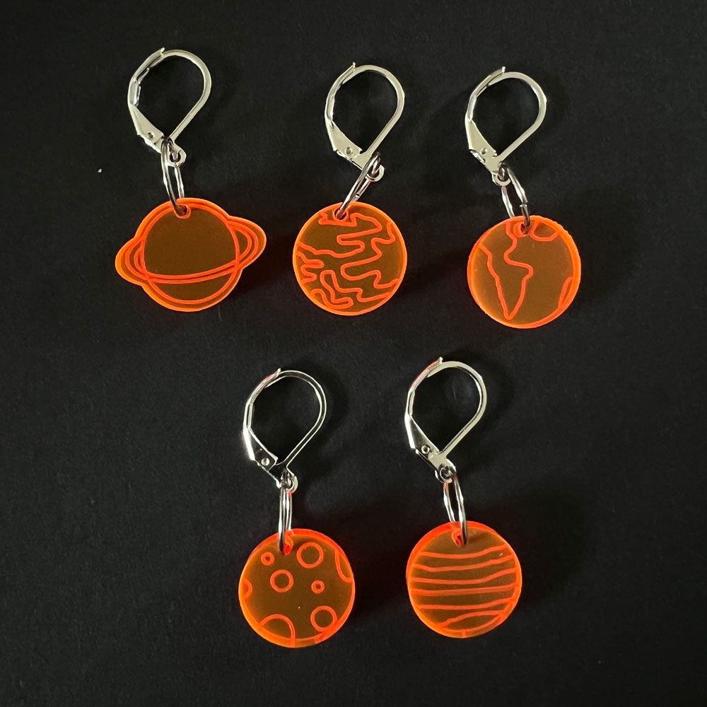 Five orange perspex stitch markers with planetary designs, each featuring a latch-back hook for easy attachment to knitting projects. Displayed on a black background.