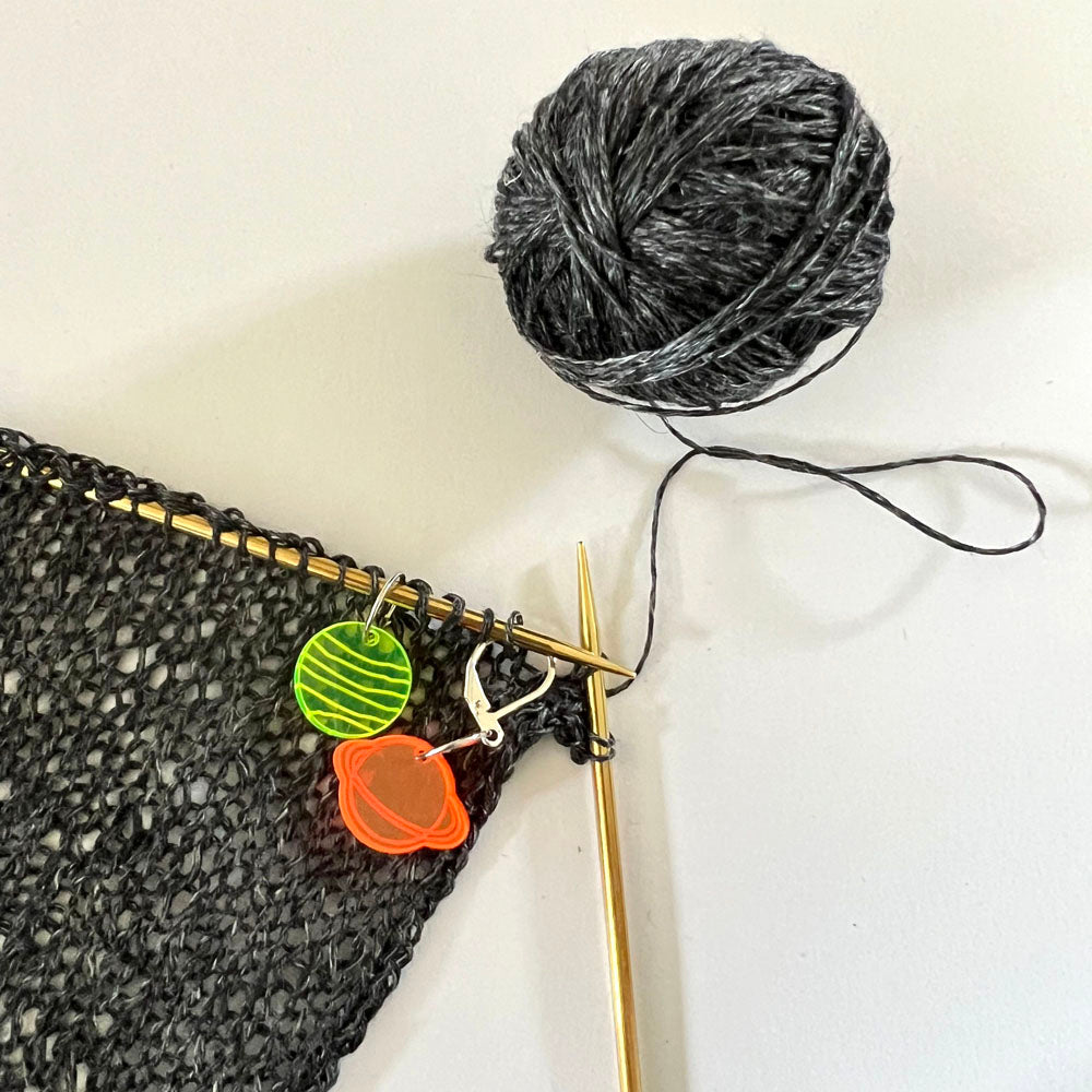 A knitting project using black yarn with two perspex stitch markers attached to the knitting needles. One marker is green with a planetary design, and the other is orange. A ball of black yarn is visible in the background.
