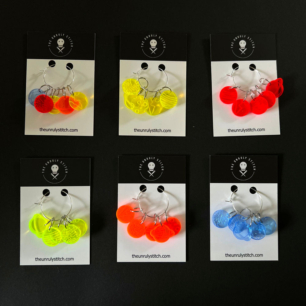 Six sets of perspex stitch markers in various colors and designs, each set displayed on a branded card from "The Unruly Stitch." The markers are arranged in a circular pattern on each card and include a variety of planetary designs.