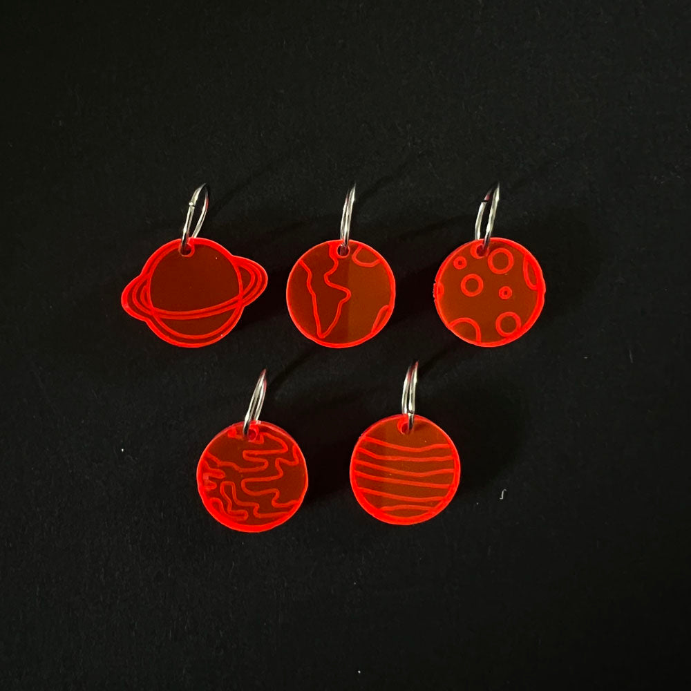 Five red perspex stitch markers featuring designs of planets and celestial bodies. Each marker is attached to a metal jump ring and arranged on a black background.