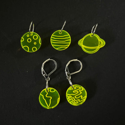 Five yellow perspex stitch markers with planetary designs, each featuring a mix of jump rings and latch-back hooks. The markers are displayed on a black background.