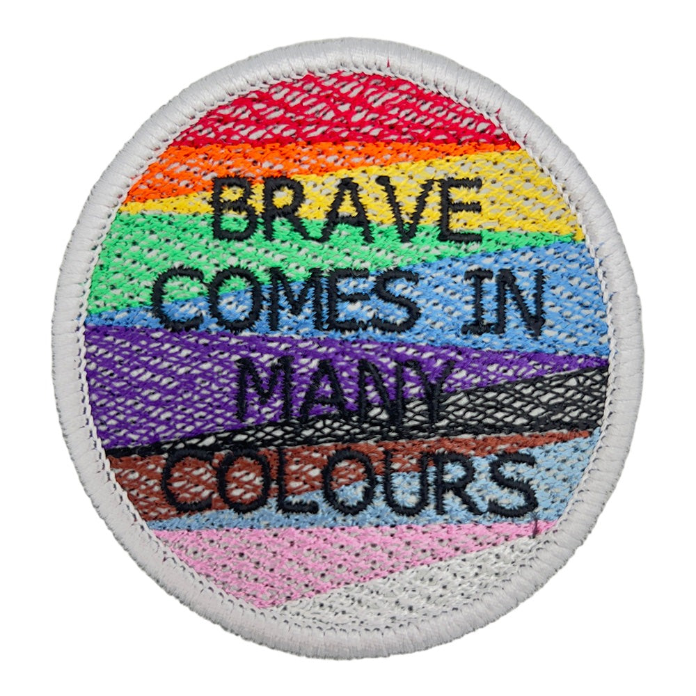 A close-up image of a circular embroidered patch with the text "BRAVE COMES IN MANY COLOURS." The patch features horizontal stripes in the colors of the rainbow pride flag, including red, orange, yellow, green, blue, and purple, with black and brown stripes.