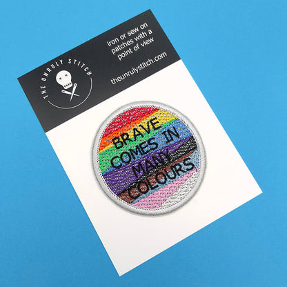 An embroidered patch with the text "BRAVE COMES IN MANY COLOURS" displayed on a white card with a blue background. The patch features horizontal stripes in rainbow colors with black and brown stripes, symbolizing pride and diversity.