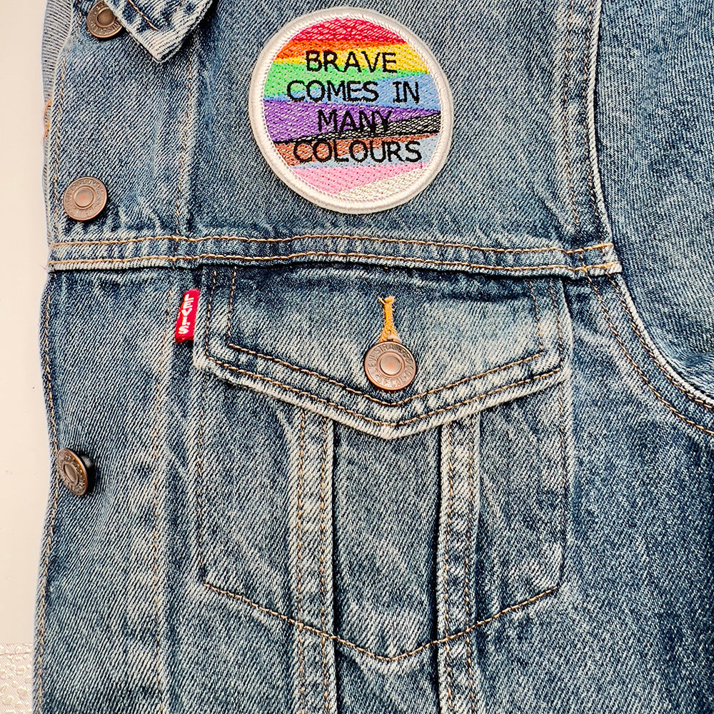 Patch on Denim Jacket: An embroidered patch with the text "BRAVE COMES IN MANY COLOURS" ironed onto the upper left section above the pocket of a denim jacket. The patch features horizontal stripes in rainbow colors with black and brown stripes, representing pride and inclusion.