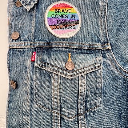Patch on Denim Jacket: An embroidered patch with the text "BRAVE COMES IN MANY COLOURS" ironed onto the upper left section above the pocket of a denim jacket. The patch features horizontal stripes in rainbow colors with black and brown stripes, representing pride and inclusion.