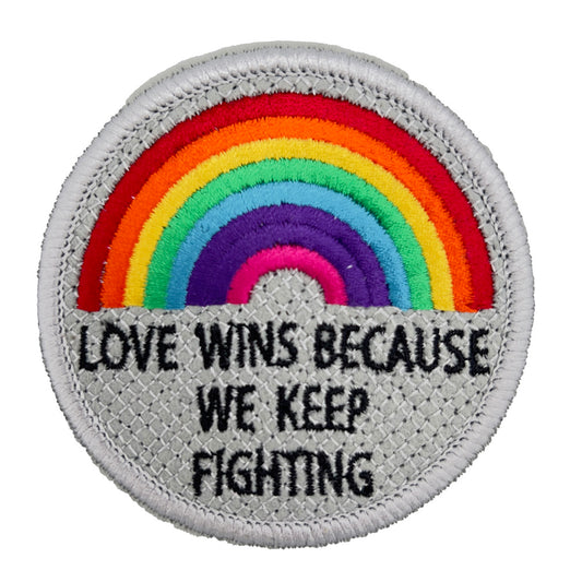 A circular embroidered patch featuring a rainbow with the text "LOVE WINS BECAUSE WE KEEP FIGHTING" below it. The background is silver with a crisscross pattern, and the border is also silver