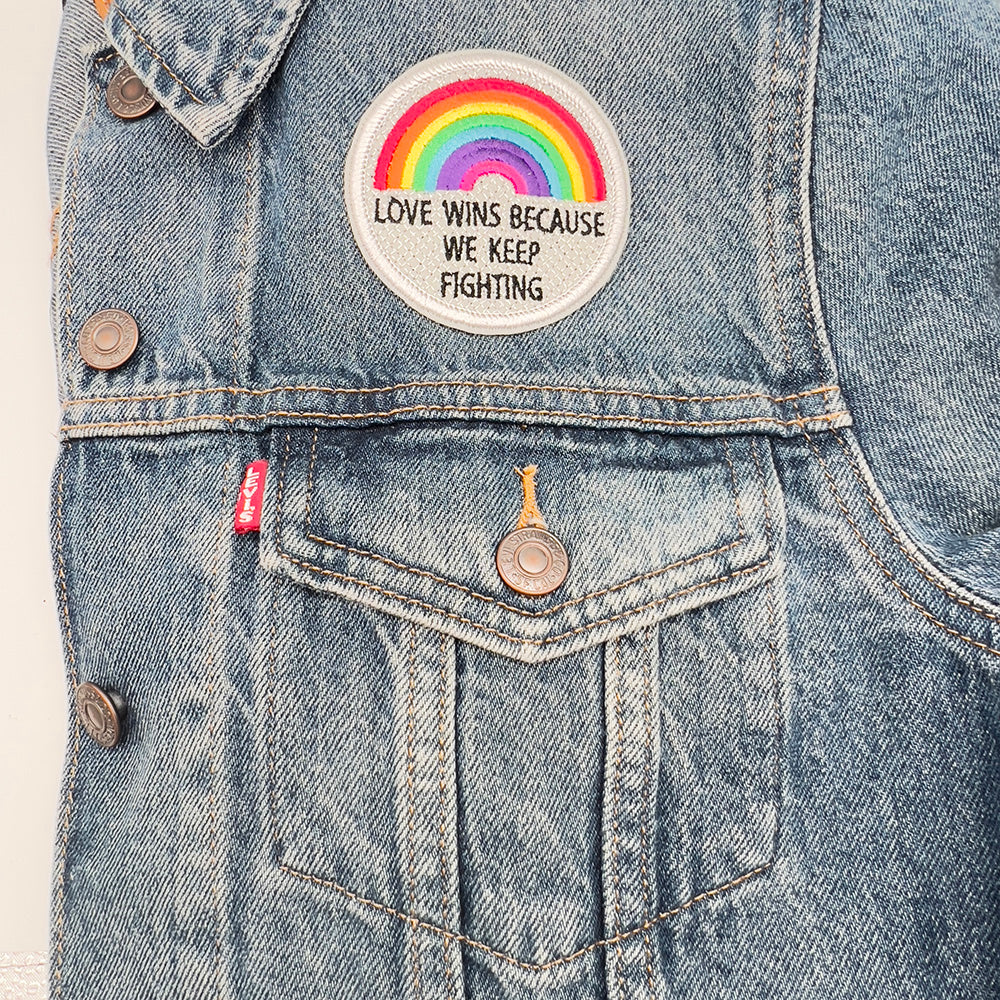 The circular embroidered patch is sewn onto the upper left side of a denim jacket. The patch stands out with its vibrant rainbow colors and pale grey background against the blue denim.