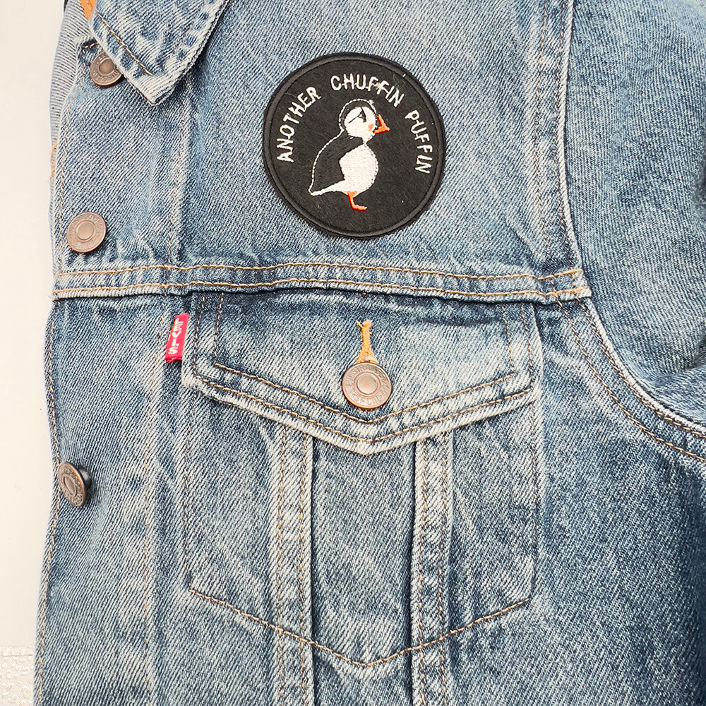A circular embroidered felt patch with a puffin design and the text "ANOTHER CHUFFIN PUFFIN" in white stitching, ironed onto a denim jacket above the pocket. The patch stands out against the light blue denim fabric.