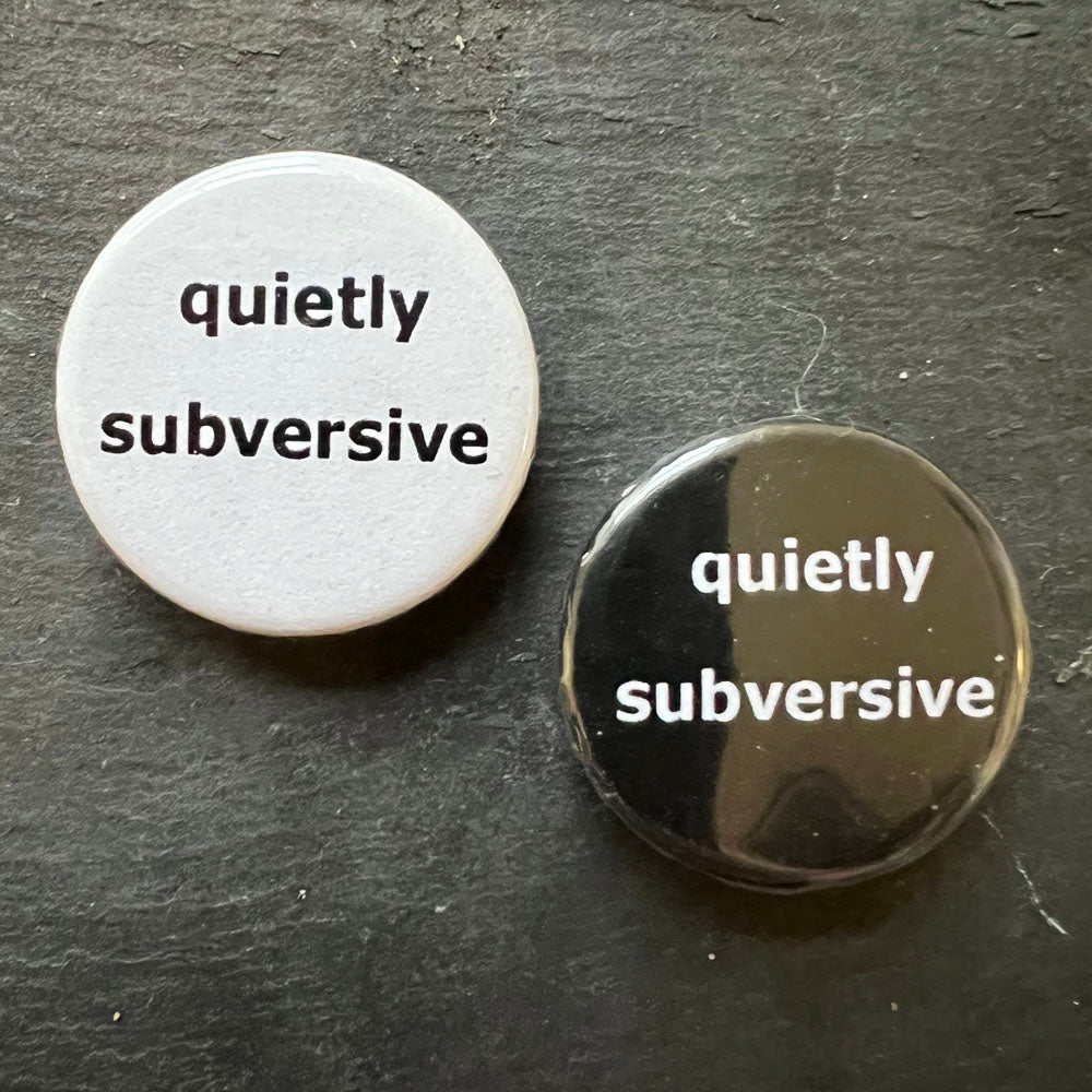 Two circular pin badges side by side on a dark surface. The badge on the left is white with black text that reads "embrace bewilderment." The badge on the right is black with white text that reads "quietly subversive".