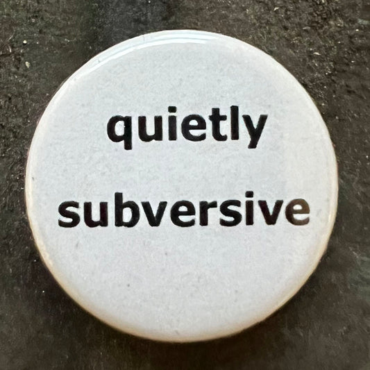 Close-up of a white circular pin badge with black text that reads "quietly subversive" against a dark background.