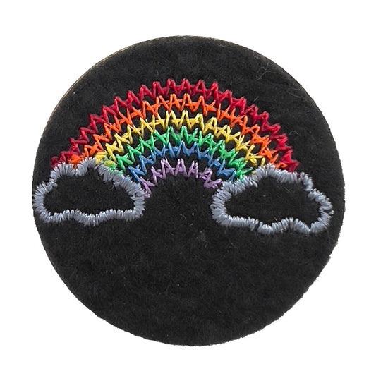 Close-up of a felt badge with an embroidered rainbow between two clouds in multicolored threads on a black background, designed by The Unruly Stitch.