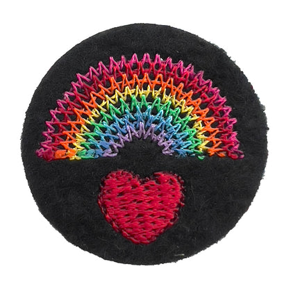 Close-up of a felt badge with an embroidered rainbow over a red heart in multicolored threads on a black background, designed by The Unruly Stitch