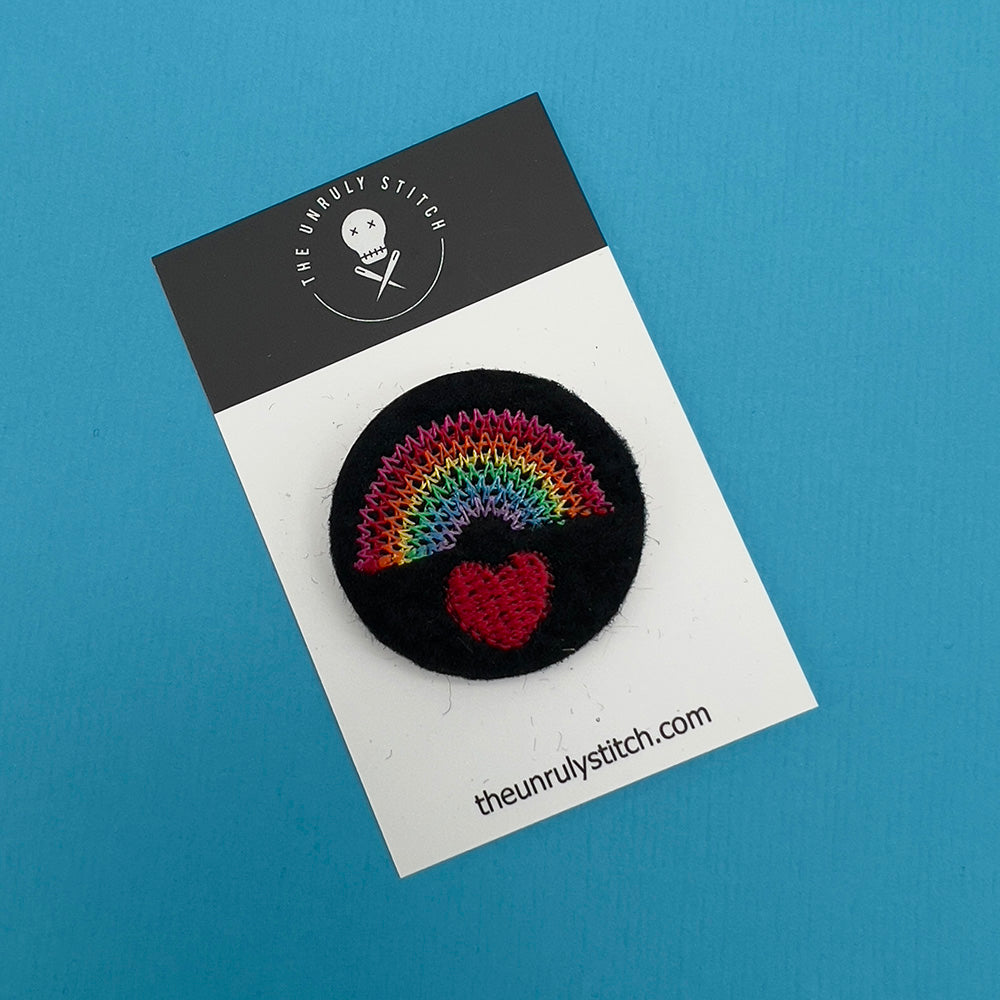 Embroidered felt badge depicting a rainbow over a red heart in multicolored threads on a black background, mounted on a branded card from The Unruly Stitch.