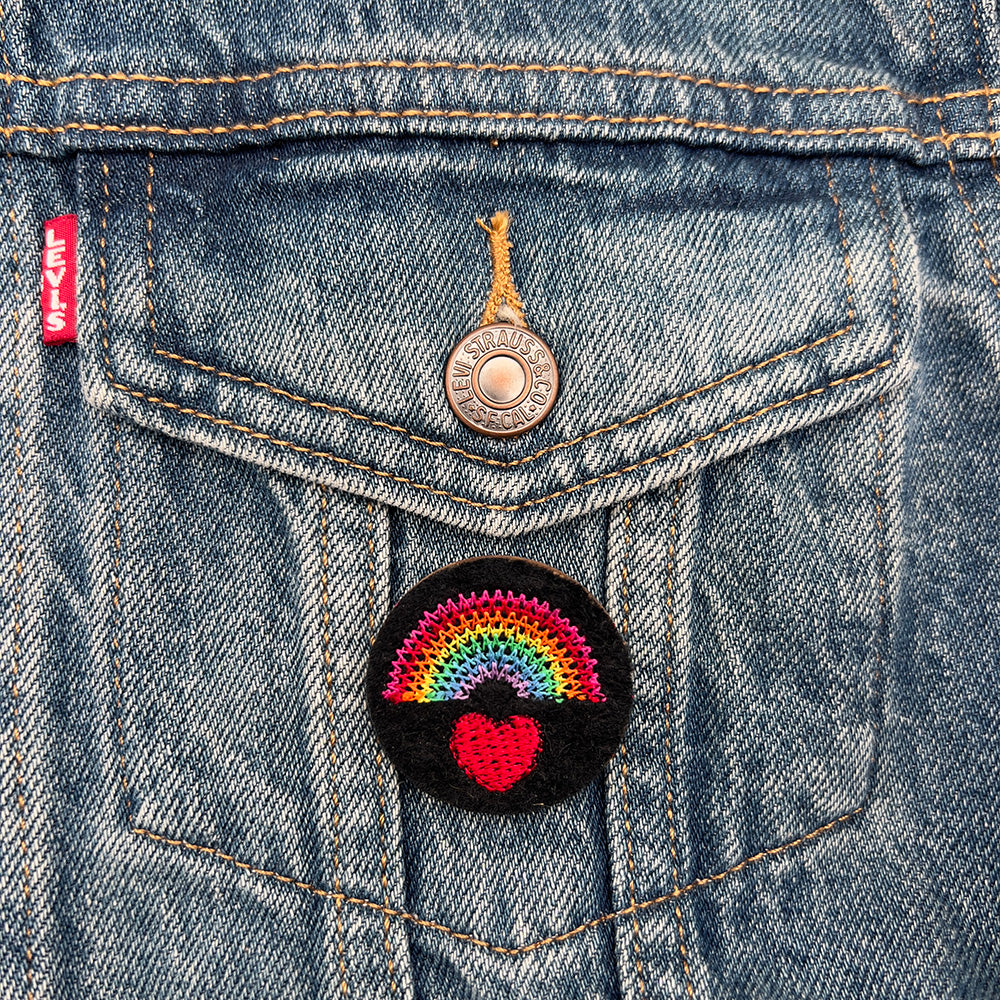 Embroidered felt badge with a rainbow over a red heart design in multicolored threads on a black background, pinned on the pocket of a denim jacket, designed by The Unruly Stitch.