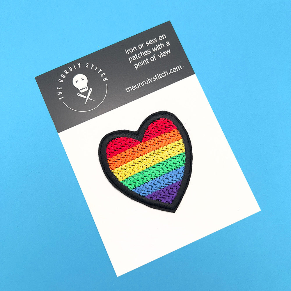 Heart-shaped rainbow pride flag patch on a white card with "The Unruly Stitch" branding, set against a blue background.