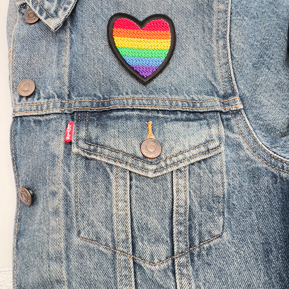 Heart-shaped rainbow pride flag patch ironed onto a denim jacket above the left-side pocket.