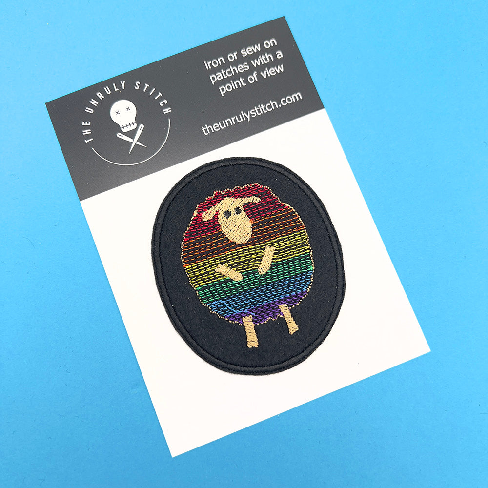 An embroidered felt patch featuring a sheep with a rainbow-colored body, outlined in beige, placed on a white card with the text "The Unruly Stitch" at the top. The card has a white background and sits on a blue surface.