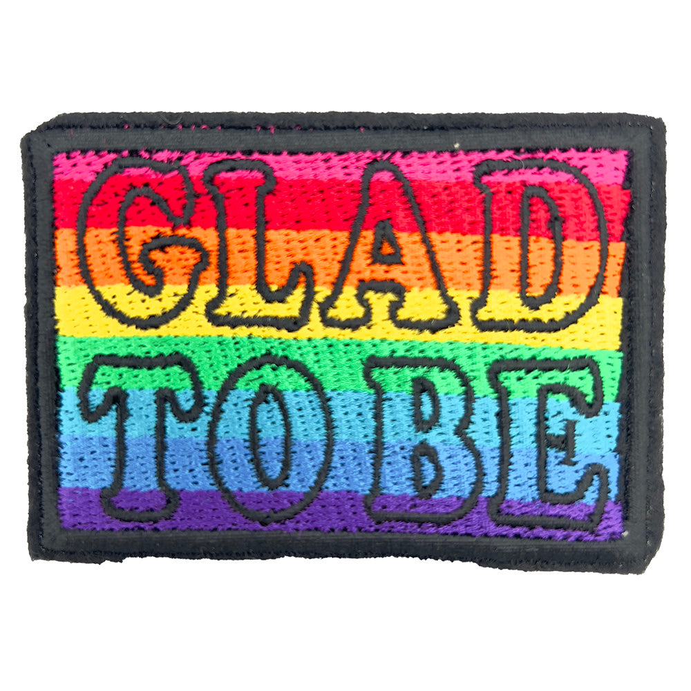 Close-up view of an embroidered felt patch with the words "GLAD TO BE" in bold black outline, filled with rainbow colors ranging from red, orange, yellow, green, blue, and purple.