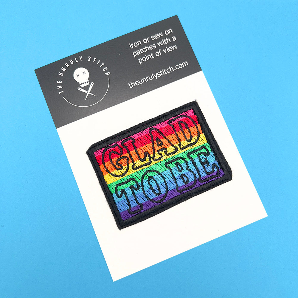  Embroidered felt patch with the words "GLAD TO BE" on a card. The patch has a rainbow gradient background with text outlined in black. The card features the branding "The Unruly Stitch" with a skull and crossed needles logo at the top.