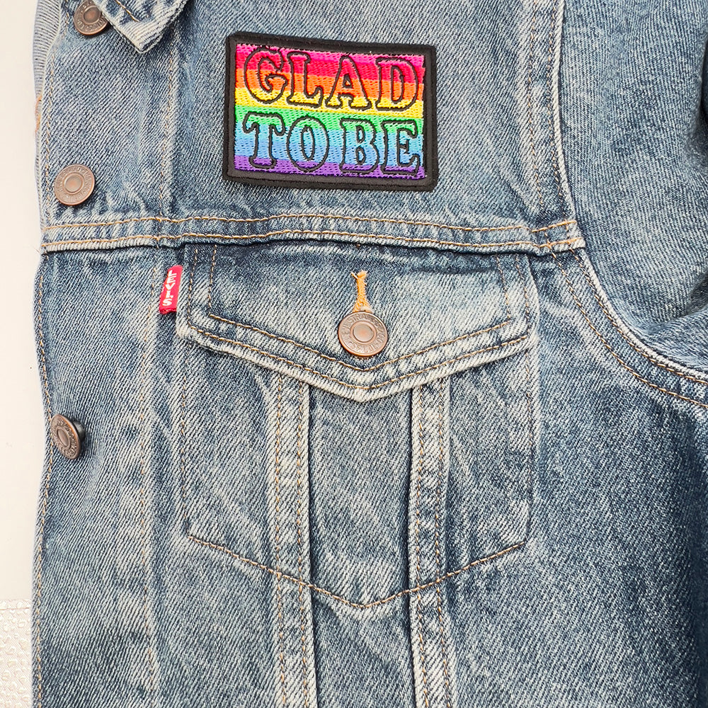 Embroidered felt patch with the words "GLAD TO BE" on a denim jacket. The patch has a rainbow gradient background with text outlined in black, shown above the pocket of a denim jacket