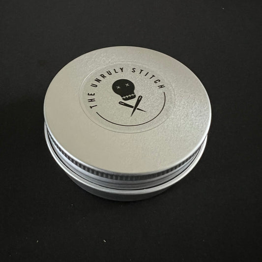 A single round metal tin with 'The Unruly Stitch' logo on the lid, closed and placed on a black background.