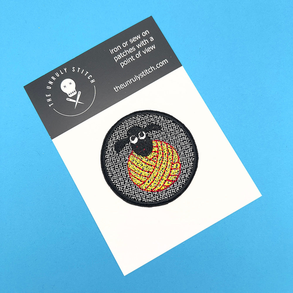 The same embroidered felt patch with the black sheep's head on a yellow and red yarn ball, displayed on a card. The card has a blue background and the brand "The Unruly Stitch" with a skull and crossed needles logo.