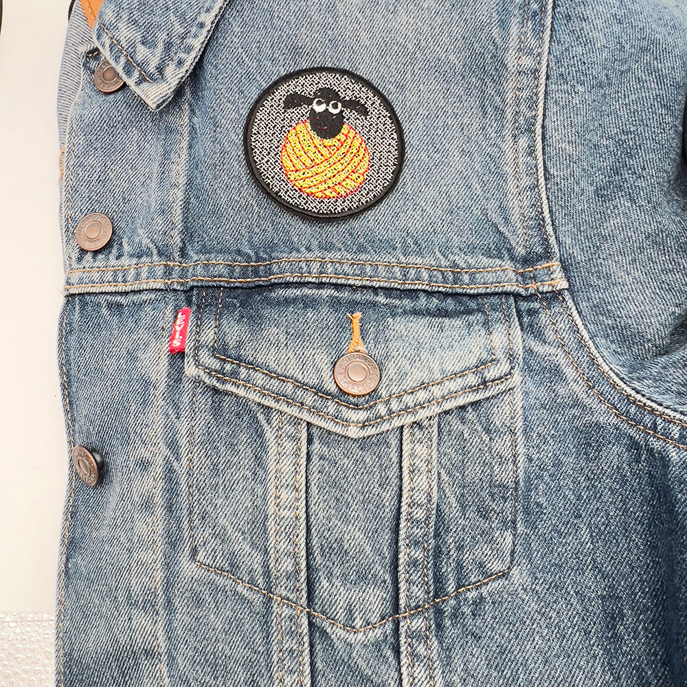 The same embroidered patch featuring the black sheep's head on a yellow and red yarn ball, now ironed or sewn onto the upper left part of a light blue denim jacket. The jacket has visible seams and buttons, emphasizing the patch's placement.