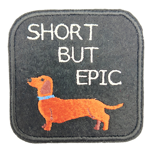 Close-up image: Close-up view of an embroidered felt patch with the text "SHORT BUT EPIC" above an image of a dachshund. The dachshund is depicted in brown with a blue collar, and the text is in white.