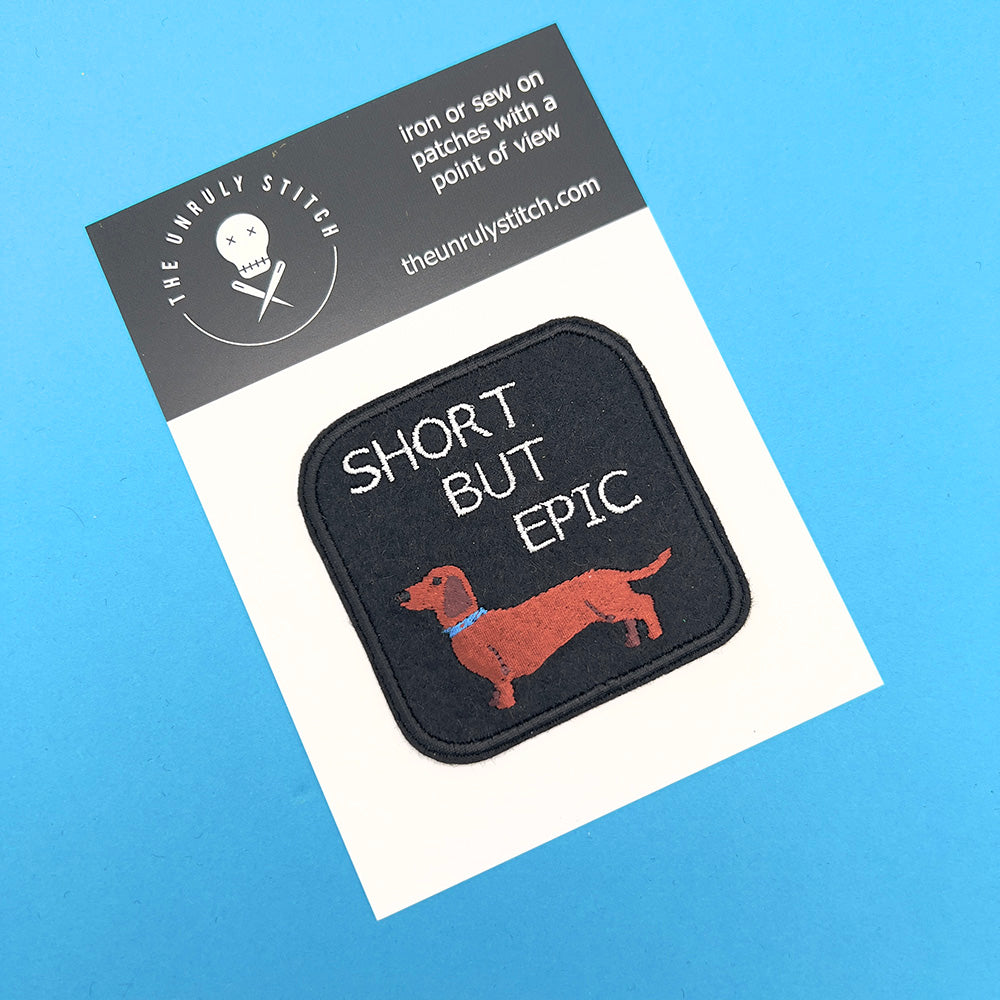 On card image: An embroidered felt patch with the text "SHORT BUT EPIC" above an image of a dachshund, displayed on a card. The dachshund is brown with a blue collar, and the text is white.