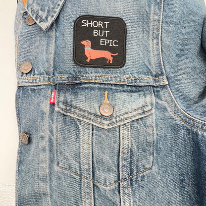 An embroidered felt patch with the text "SHORT BUT EPIC" above an image of a dachshund, attached to the front of a denim jacket. The dachshund is brown with a blue collar, and the text is white.